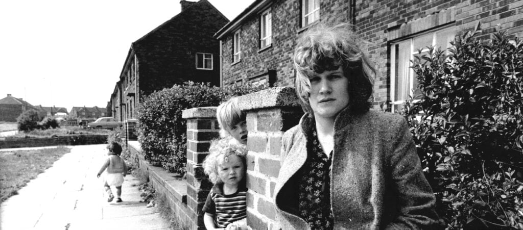 Andrea Dunbar, black and white photo of a woman with hair down to her shoulders, outside a house with two children running around behind her.