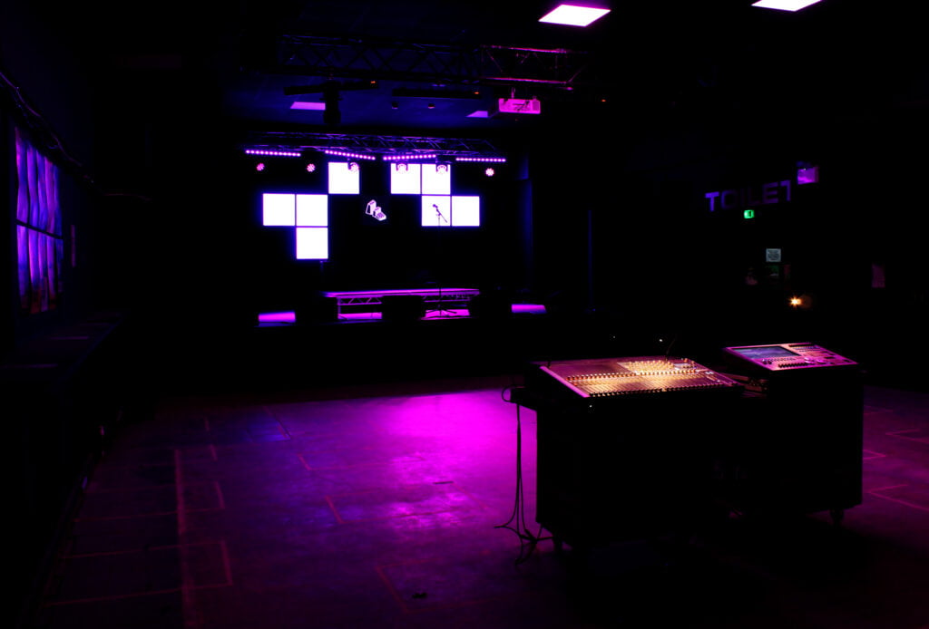Dark interior with purple light on the floor, coming from a stage visible in the background
