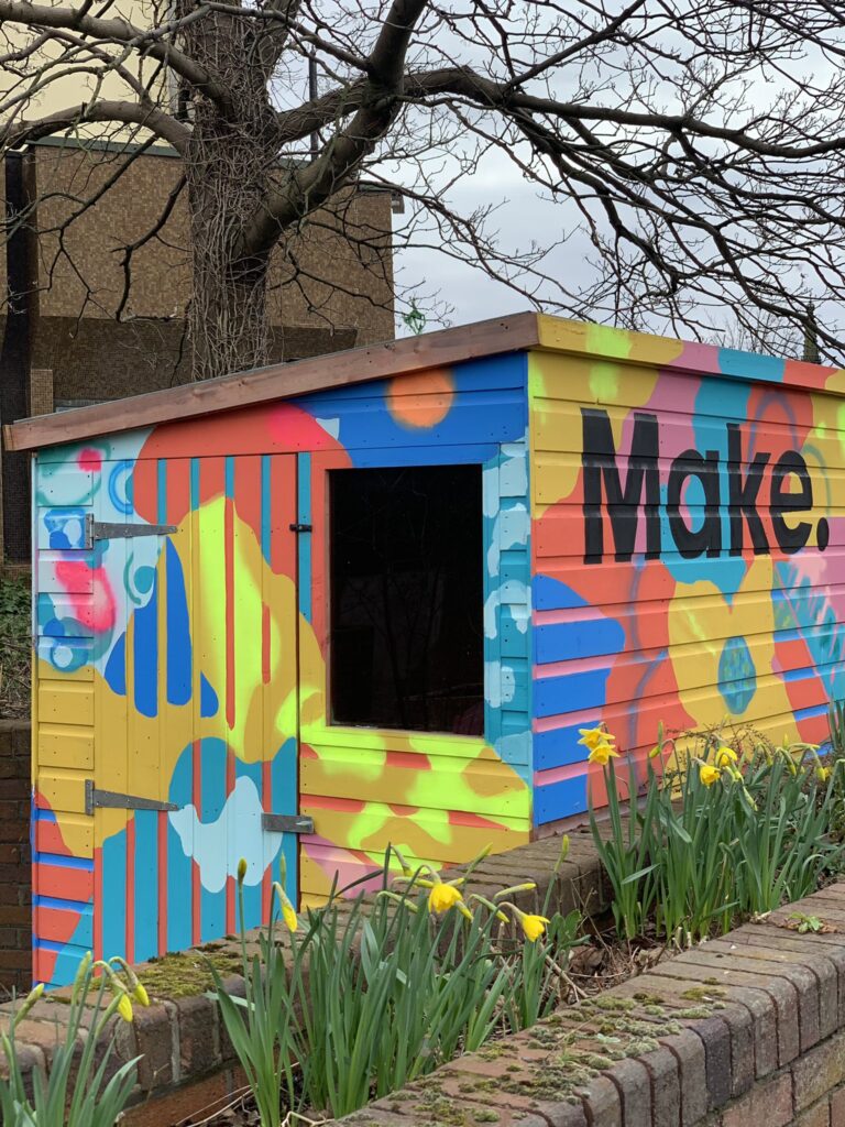 Brightly coloured painted shed with floral motifs and the word 'Make.' painted on the right hand side. Daffodils in the foreground and a leafless tree in the background.