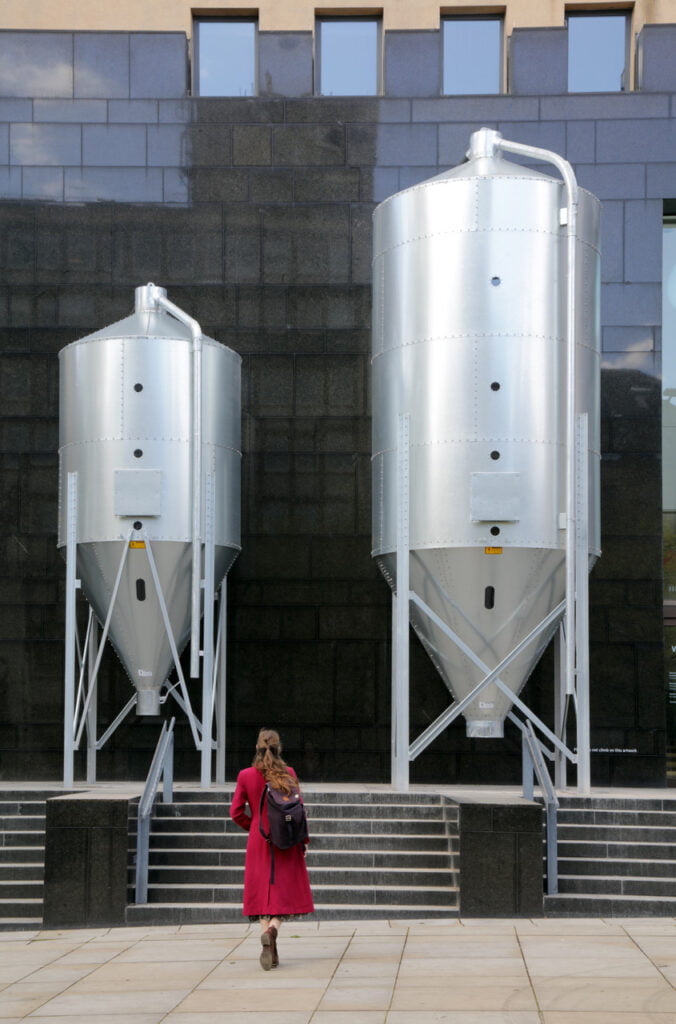 Two large silver grain silos in front of a reflective, black wall at the front of a building. At the bottom, we see the back of a woman, with long brown hair, in a raspberry pink coat, walking towards them.