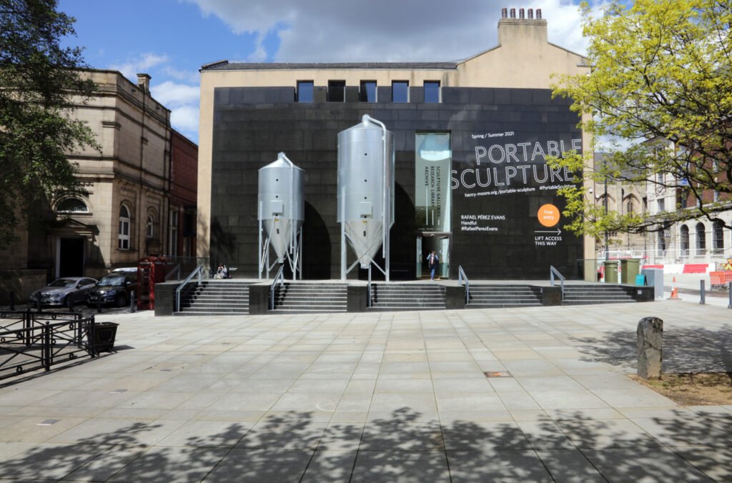 The front of the Henry More institute with a big black wall with the words 'Portable Sculpture' in big letters, next to two large, silver grain silos on silver legs. Below is a concrete staircase and a green tree on the right, in the sun. 