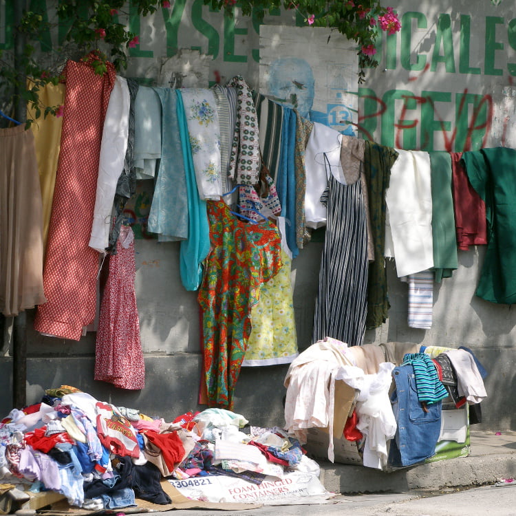 A wall of clothes hanging on a line, with piles on the ground and in a box. There is grafitti on the wall.