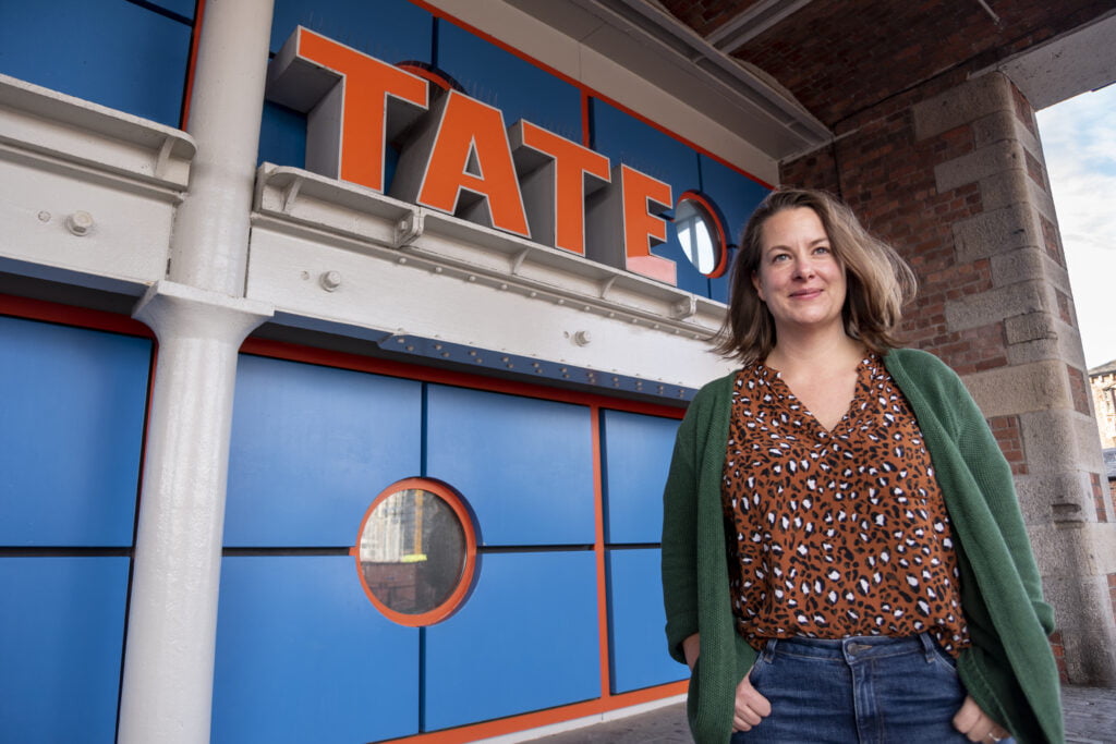 Portrait of Emily Speed outside the Tate Liverpool sign at the entrance. She is a white woman with medium length dark blond hair, wearing an animal print brown top, green cardigan and jeans