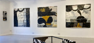 Three large square paintings by artist Christopher Tansey. Layered, rounded shapes of grey, white, yellow and black.