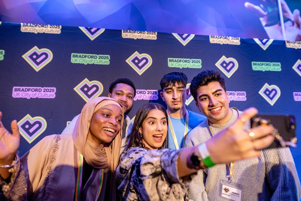 Photo of a group of young people taking a selfie in front of a Bradford 2025 poster. 