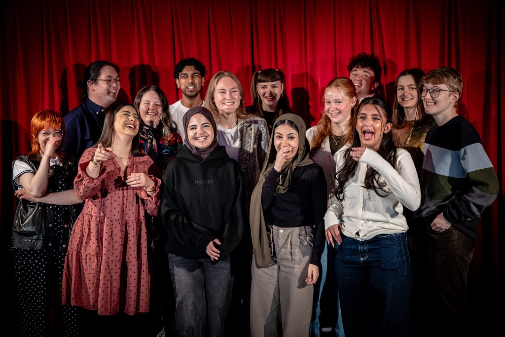 Photo of a group of young people stood in front of a red curtain laughing. 