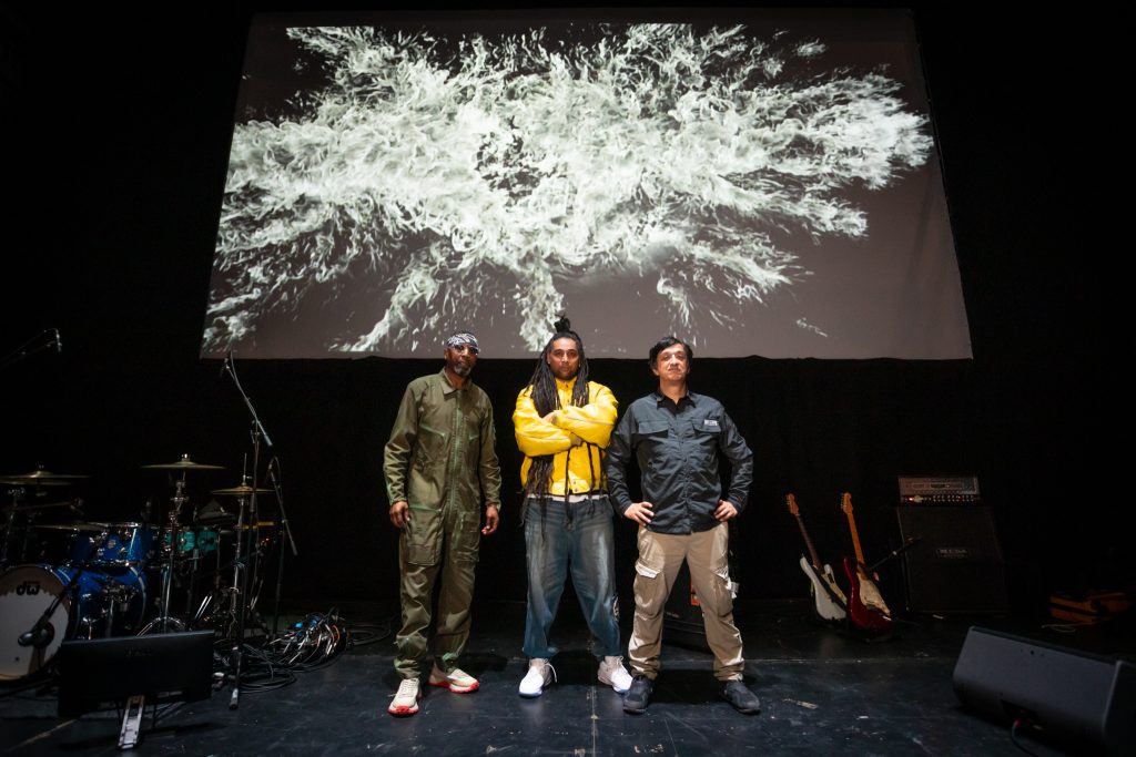 A photo of Asian Dub Foundation stood on stage in front of a screen