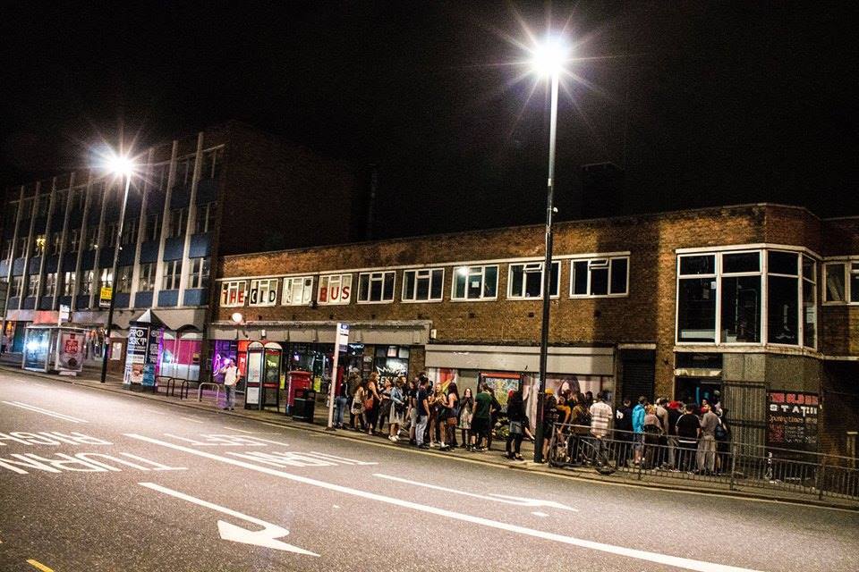 A photo of a nightclub from across the road with people stood outside.